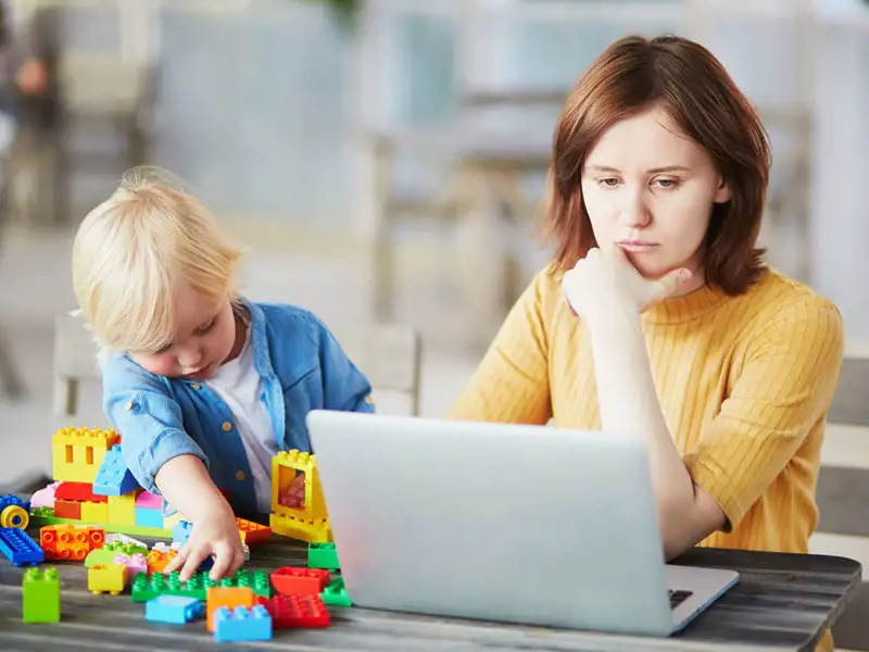 Woman sitting at a table on her laptop while her son plays with blocks