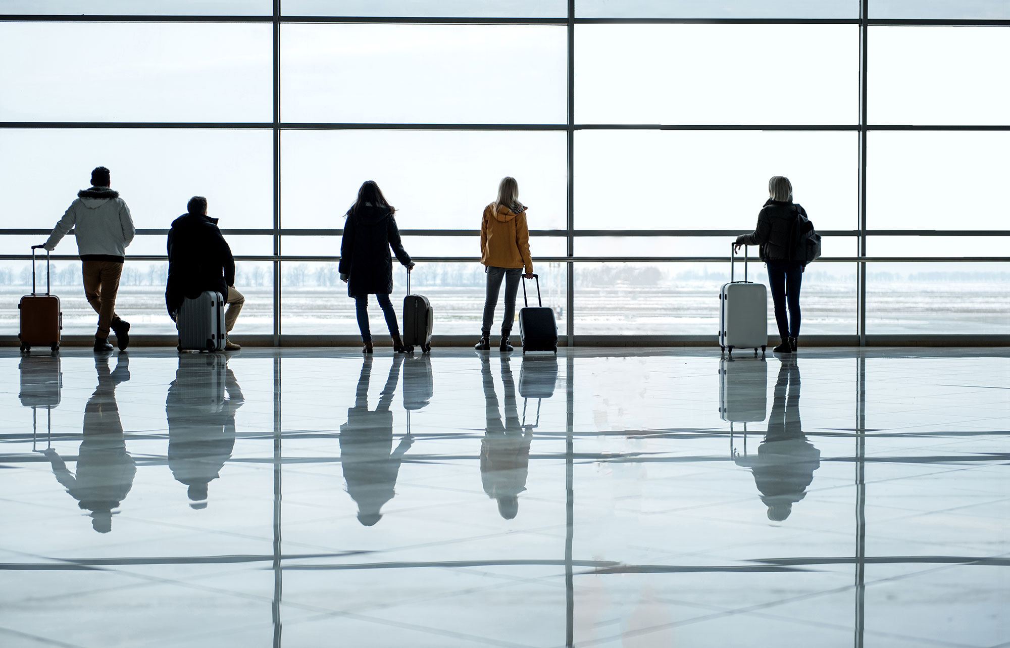 Airport patrons stand near a window with their luggage.